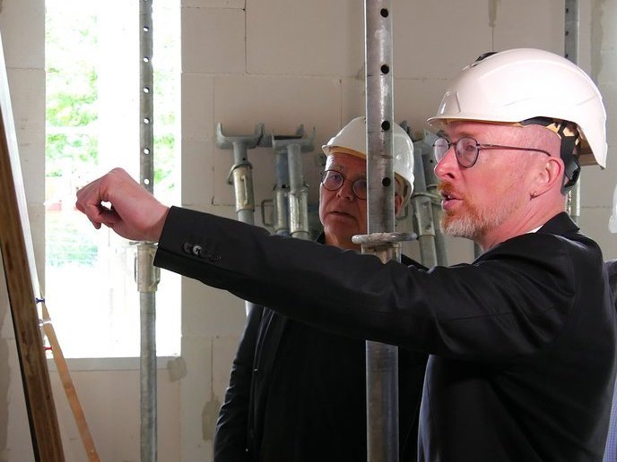 Person im schwarzen Anzug zeigt auf etwas, eine weiter beobachtet ihn, sie stehen in der Baustelle