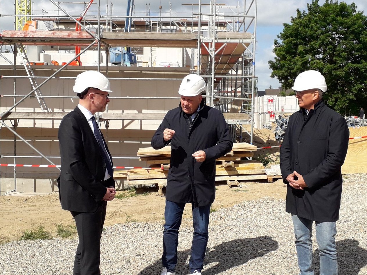 drei Personen stehen auf einer Baustelle, Die Person in der Mitte erklärt etwas. Links trägt Anzug, Mitte und rechts schwarze Jacke und Jeans