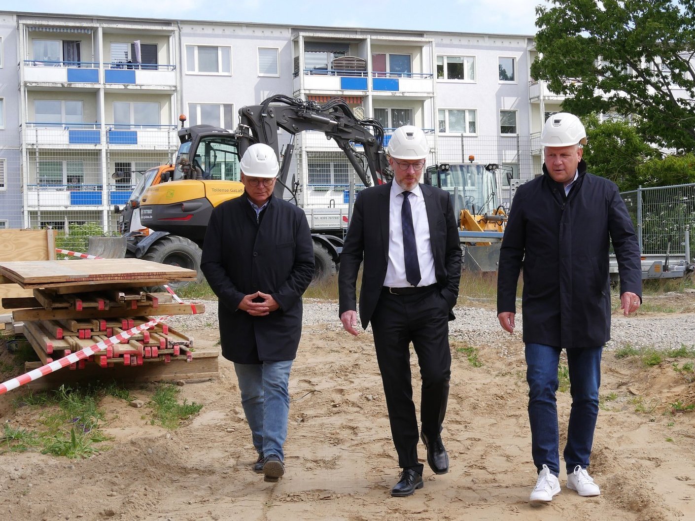 drei Personen gehen über einer Baustelle, Mitte trägt Anzug, links und rechts schwarze Jacke und Jeans