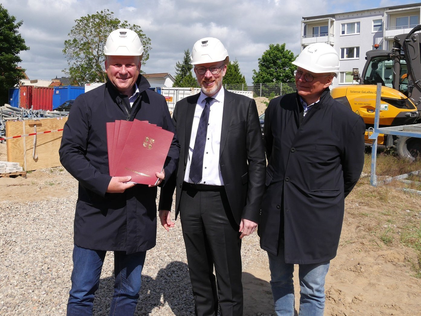 Drei Personen lächeln in die Kamera, Links hält Unterlagen in den Händen, sie stehen auf der Baustelle